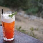 A vibrant citrus cocktail in a crystal glass with a rosemary garnish on an outdoor table.